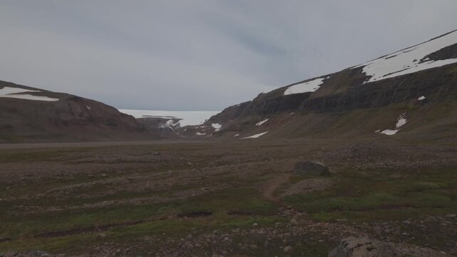 Remote glacial valley with rocky tundra, lingering snowfields and Drangajokull ice cap under overcast sky, static establishing, nobody
