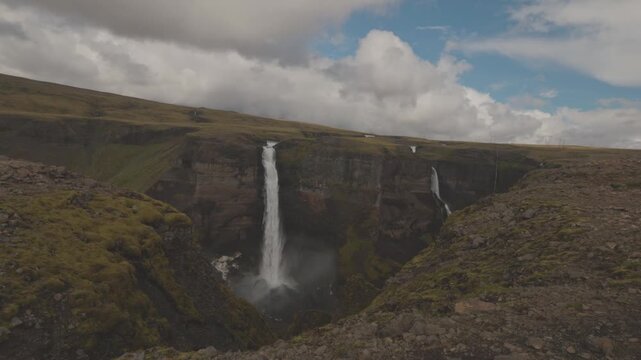 Haifoss plunges from a high plateau into a steep rocky canyon beneath shifting clouds in Iceland, establishing handheld static