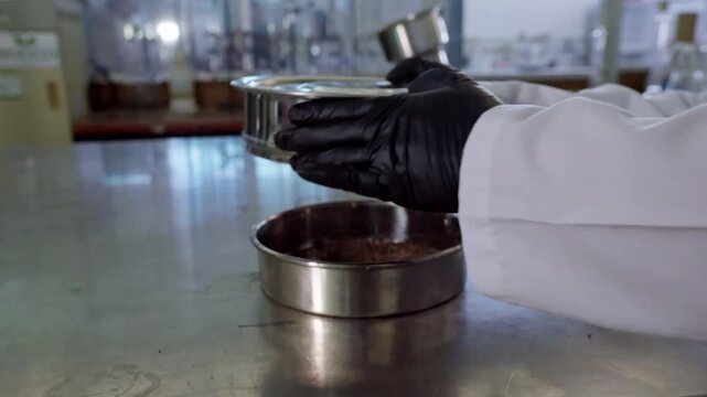 Close-up of a scientist wearing black gloves and a lab coat, sifting a soil sample through a sieve in a laboratory. This process is crucial for agricultural analysis.