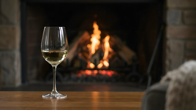 A glass of white wine sitting on a wooden table in front of a cozy, glowing fireplace