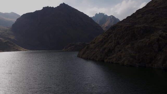 Low aerial shot gliding over the deep rippling waters of an alpine glacial lake surrounded by massive, shadowy mountain peaks in the Peruvian Andes, Peru.