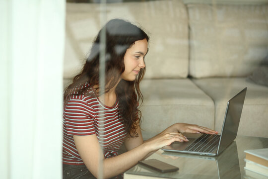 Young woman using laptop at home viewed through a window