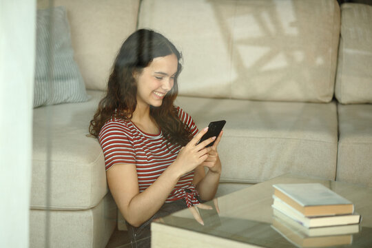 Happy teen using phone at home viewed through a window