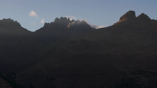 Cinematic aerial view of dramatic, shadowy Andean mountain peaks and jagged ridges glowing under the early morning sunrise light and blue skies in Cusco, Peru.