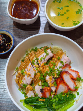 A bowl of dry egg noodles topped with crispy roast pork belly, char siu, greens, fried garlic, and spring onions, served with clear soup and dipping sauces.