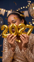 Fototapeta premium Woman holding golden 2027 number balloons while wearing a sparkling sequin top at a festive indoor celebration
