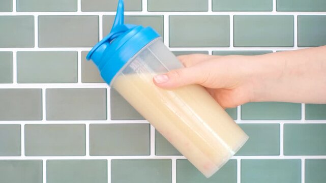 Female hand holds shaker with protein shake against a background of blue tiles.
