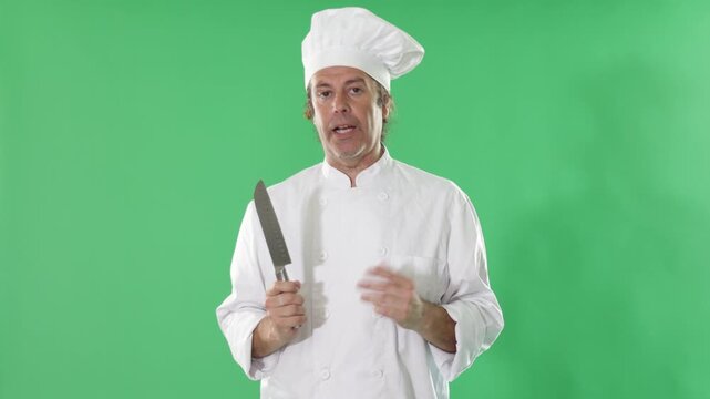 Adult male chef wearing white cook uniform and hat stands against green screen, holding chef's knife and explaining chopping or slicing method 