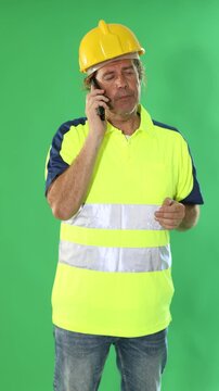 Adult male worker wearing reflective polo shirt and safety hard hat stands against green screen, holding smartphone and speaking 