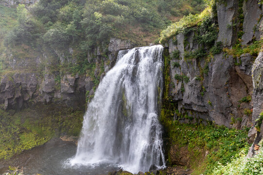 Scenic view of Snow Leopard waterfall flowing over a rugged rock cliff into a calm pool, surrounded by lush green forest vegetation and mossy stone walls
