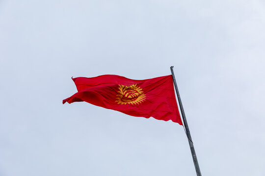 Close-up of the red Kyrgyzstan flag with yellow sun and tunduk symbol flying on a tall flagpole against an overcast sky, patriotic national identity concept