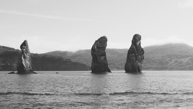 Black and white seascape with the Three Brothers sea stacks rising from Avacha Bay, Kamchatka Krai, Russia, with calm water, distant hills and cloudy sky