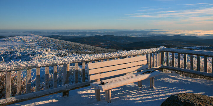 A snow-covered wooden bench and railing offering a breathtaking view of a vast mountain range under a clear blue sky. The sun casts long shadows across the icy landscape.