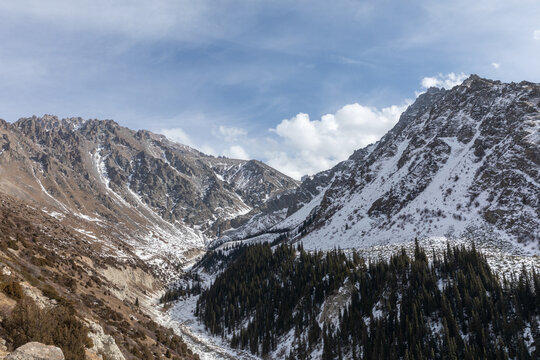 Scenic winter view of rugged Tian Shan mountains and pine forest in Ala Archa National Park, Bishkek region, Kyrgyzstan, under blue sky with clouds
