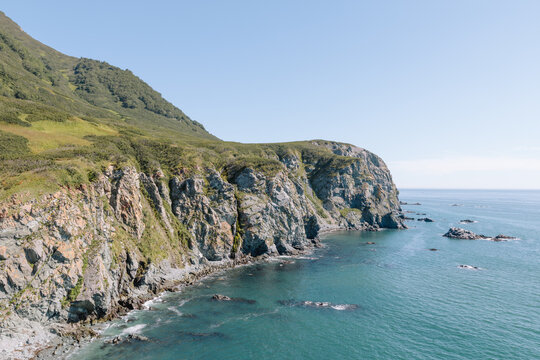 Scenic rocky coastline with steep sea cliffs, green slopes and calm blue water under clear sky, summer seascape on Kamchatka Peninsula, Russia