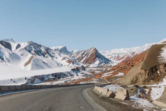 Wide view of the Pamir Highway with a winding asphalt road through snow covered mountains and red hills under clear blue sky in Kyrgyzstan, Central Asia