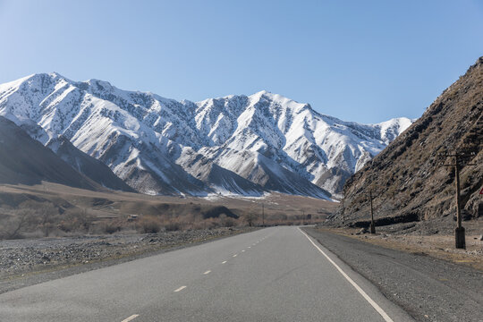 Empty asphalt road on the Pamir Highway leading through a high mountain valley with snow covered peaks under clear blue sky, Kyrgyzstan, Central Asia