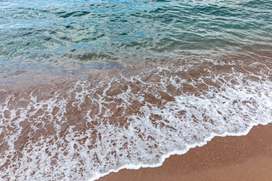 Close-up view of small ocean waves and white foam washing onto a sandy beach, with clear turquoise water and natural textures, ideal for summer travel backgrounds