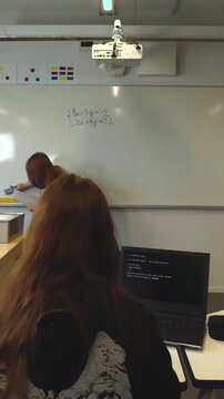 Rear view of a student watching her professor erasing a system of linear equations from the whiteboard during a math lesson, with a laptop showing code on the desk in the foreground