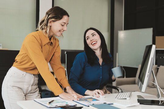 Two businesswomen discussing work using a laptop in a modern office lounge area, showing teamwork and communication