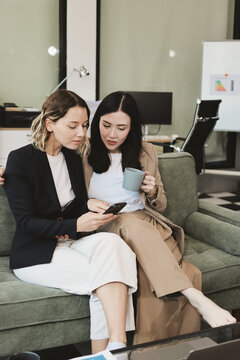 Two smiling women are comfortably sitting on a couch, sharing a moment and collaborating on a tablet, working together in a home office environment