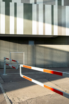 Red and white parking barrier crosses a concrete service entrance beneath striped facade panels, creating a graphic urban infrastructure detail