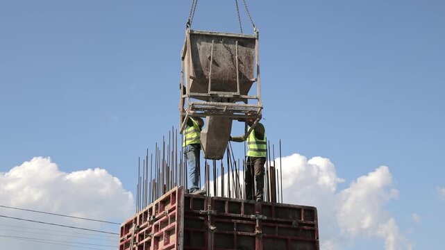 Construction workers guide suspended concrete hopper above reinforced formwork at building site, Crane lifting equipment supports active concrete pouring under clear blue sky.
