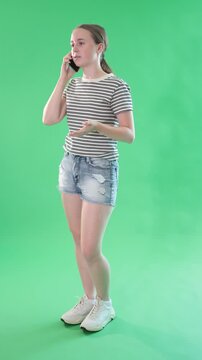Portrait of european teen girl dressed in summer stylish shorts talking by smartphone standing against green screen 
