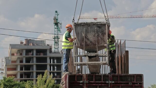 Construction workers guide concrete hopper above reinforced formwork at active building site, Tower cranes and unfinished structure frame industrial concrete work.