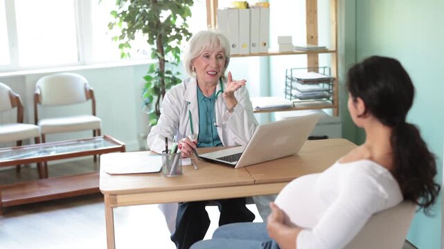 At clinic, pregnant woman is at doctors appointment. Senior medical female specialist and patient sit at table and communicate, interview, anamnesis, patient complaints 