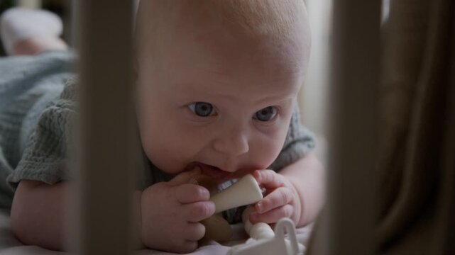 Caucasian infant lying on stomach chewing silicone teether toy inside crib. Clip suitable for parenting tutorials, infant wellness campaigns, baby product marketing