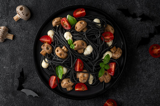 Halloween-themed food. Black squid ink spaghetti with mozzarella skulls, mushrooms, cherry tomatoes and fresh basil. Creative spooky food styling for festive autumn celebrations. Top view.