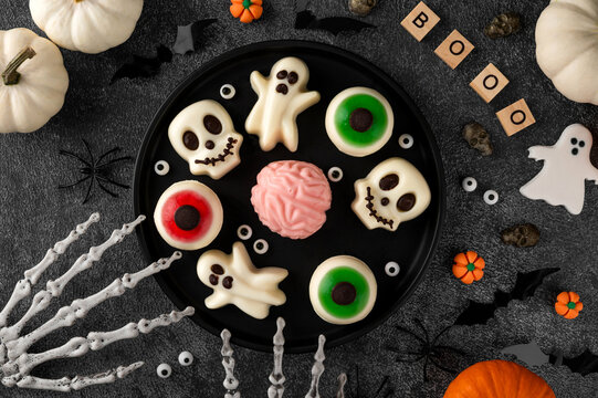 Halloween jelly desserts shaped like spooky ghosts, skulls and eyeballs, served on black plates with pumpkins and festive decorations. Top view, copy space.