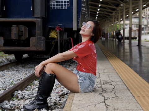 An Asian woman with short hair wearing sunglasses, a short-sleeved red vinyl top, distressed denim shorts, and black knee-high lace-up boots sitting on the edge of a railway platform