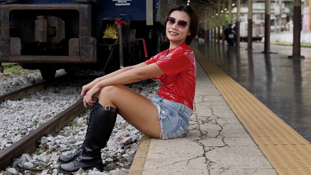 An Asian woman with short hair wearing sunglasses, a short-sleeved red vinyl top, distressed denim shorts, and black knee-high lace-up boots sitting on the edge of a railway platform