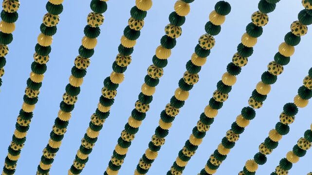 Green and yellow paper lanterns hanging for a Spanish festival against a clear blue sky, celebrating a traditional event