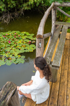 Young woman with dark hair wearing a white sweater sits on wooden dock beside a pond with lily pads and pink flowers in a serene natural setting