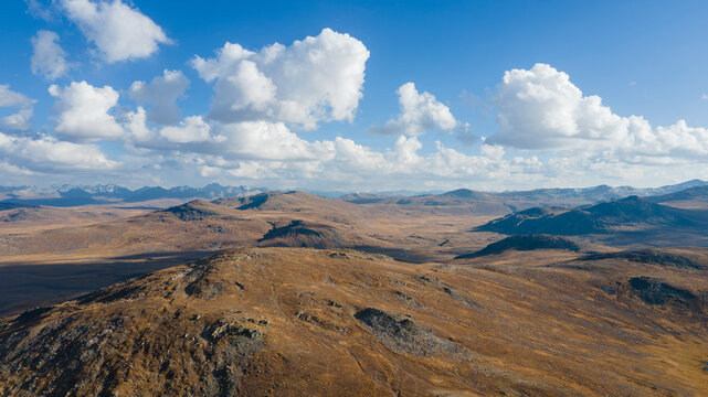 Aerial view of Deosai National Park featuring vast rolling hills, rocky terrain, and scattered clouds under a clear blue sky in Skardu, Gilgit, Pakistan.