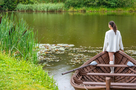 Young woman in white sweater stands on wooden boat by calm lake surrounded by lush greenery and lily pads, enjoying serene nature view in summer