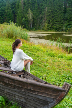 Young woman in white sweater and knee-high boots sits on an old wooden boat, surrounded by lush green grass and trees in a natural outdoor setting