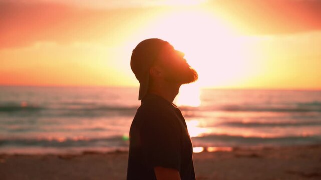 Melancholic young man with a beard and cap standing on a beach during a vibrant golden sunset, contemplating life and finding solace while looking at the sea