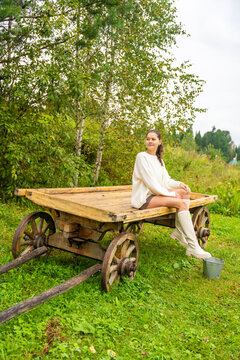 Young woman in white sweater and knee-high socks sitting on a wooden cart in a lush green field surrounded by trees and cloudy sky