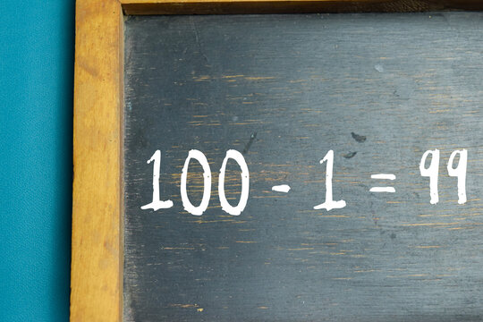 Chalkboard with phrase 100 - 1 = 99 subtraction equation on wooden desk. Basic arithmetic and school education