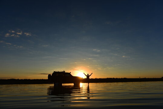 Silhouette of an adventurous man standing on an off-road car in a shallow lake during a beautiful sunset with water reflection.