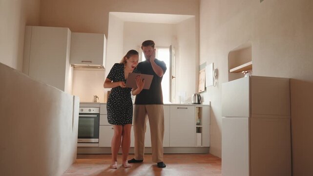 Excited young couple celebrating winning an online auction on a laptop in their new apartment kitchen, feeling happy, dancing, kissing, and giving high five