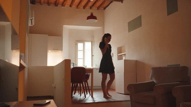 Young woman walking barefoot in her modern apartment while talking on the phone, expressing various emotions and gesturing during the conversation with natural light