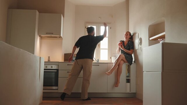 Affectionate young couple having fun, laughing, and cooking together in a bright, cozy kitchen, the cheerful woman sits on the counter while the man playfully juggles