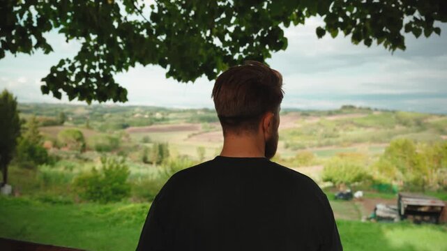 Man with a beard looking at the amazing tuscan hills from a scenic balcony with tree leaves framing the view, enjoying the nature and the peaceful italian landscape