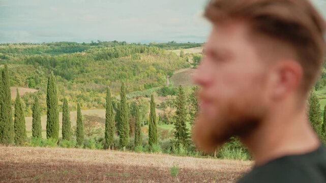 Pensive young man with a beard watching the tuscan landscape with cypress trees. Traveler finding inspiration and enjoying a peaceful moment in the italian countryside