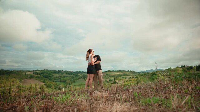 Romantic couple running, hugging, and kissing in a beautiful tuscany landscape, a woman embracing her man in the middle of a green hill with an amazing view on a cloudy day